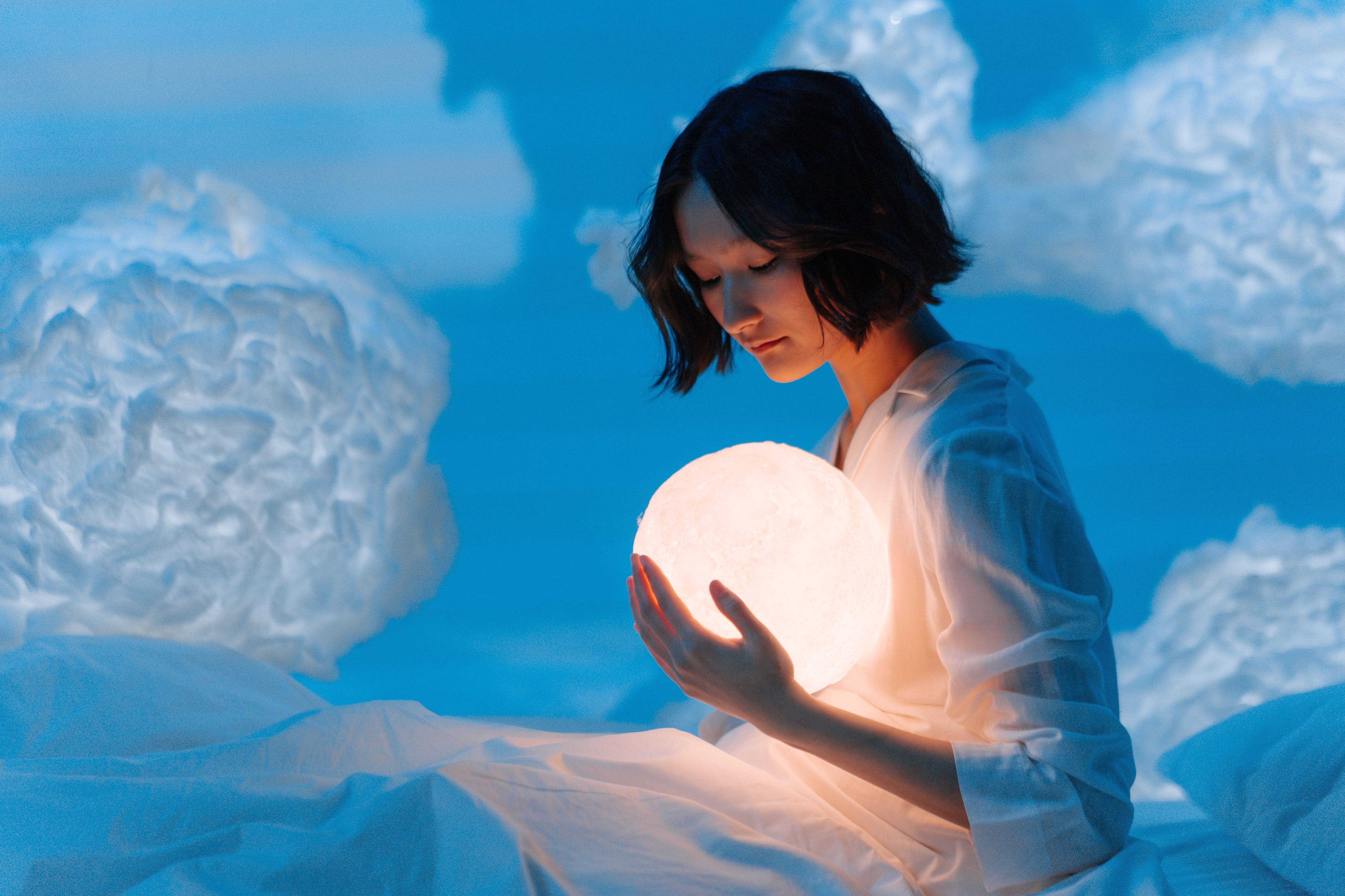 Woman holding a glowing sphere in a dreamlike cloud-filled setting, suggesting imagination and escapism.