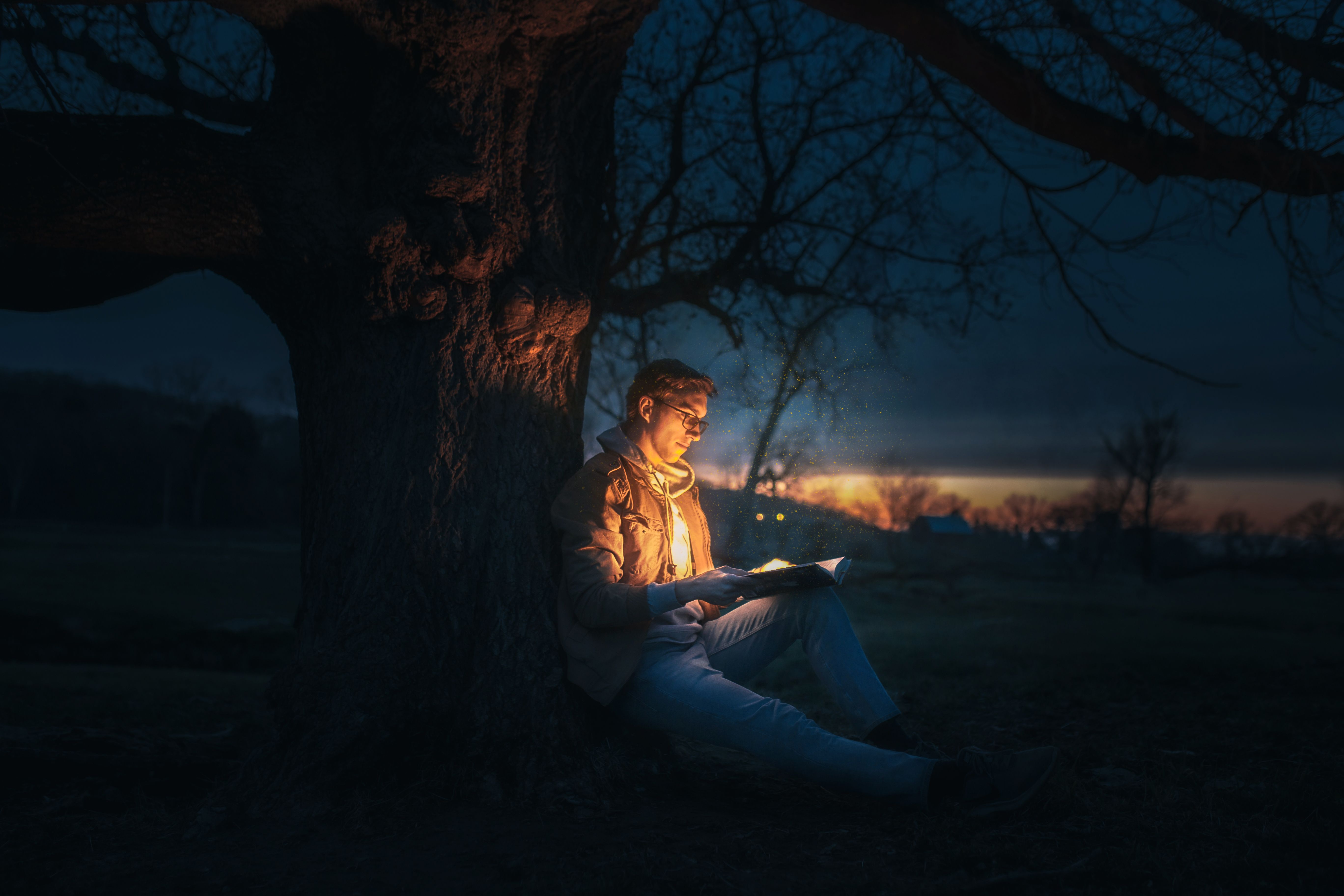 Man reading beneath a tree at night while illuminated by a glowing book, evoking fantasy and escape.