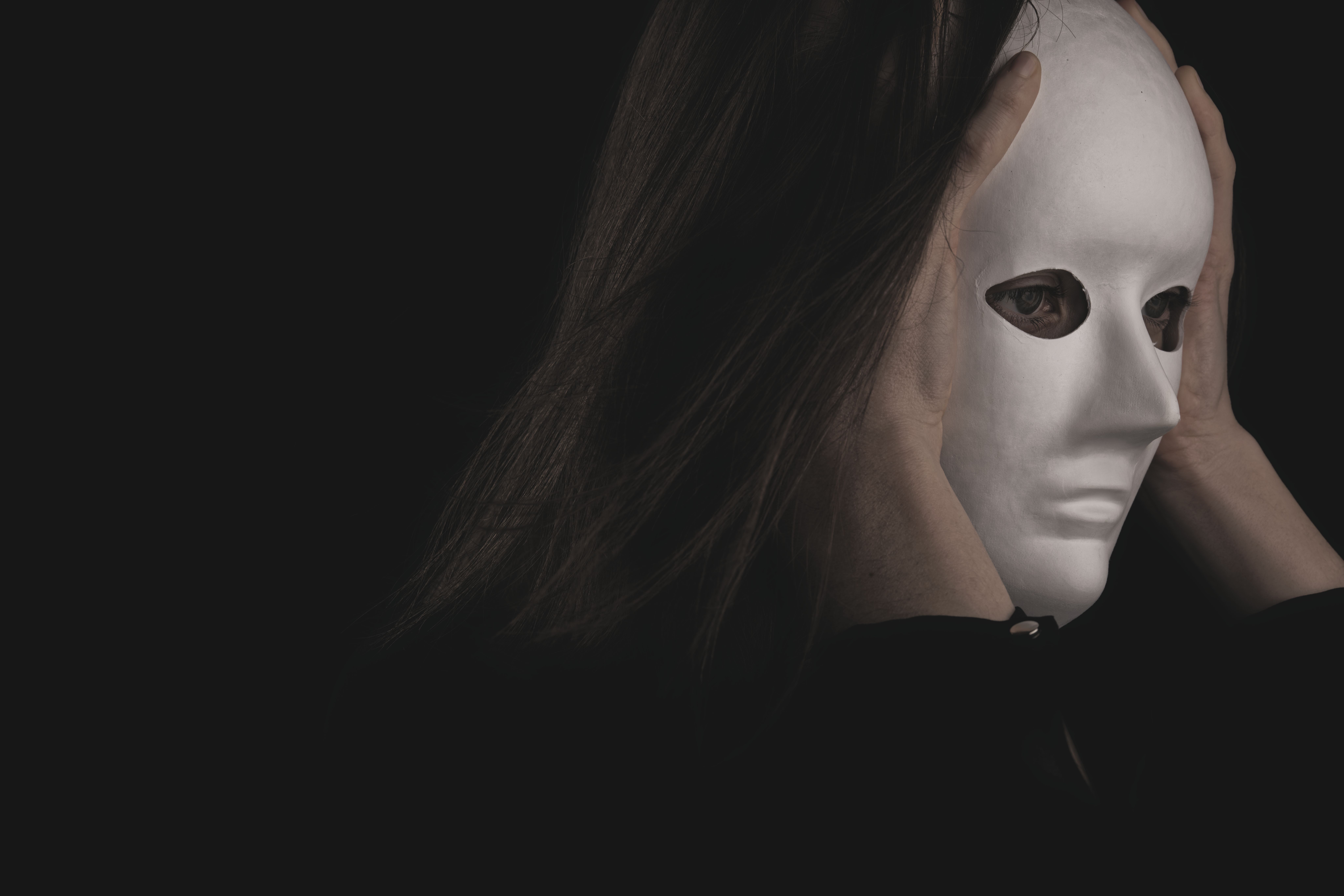 Person holding a white mask against a dark background, symbolizing identity and taking on another role.